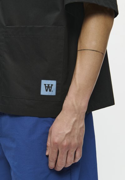 Black short-sleeve shirt with a patch pocket, featuring a light blue logo label. Worn with blue textured shorts.