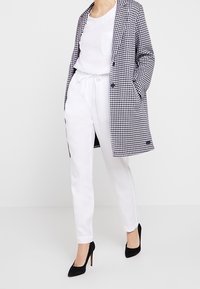 Manteau à motif vichy en noir et blanc, porté sur un haut blanc et un pantalon blanc. Fini avec des chaussures à talons noirs.