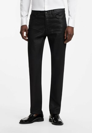 Mann trägt eng anliegende schwarze Hose, weißes Hemd und glänzende schwarze Anzugsschuhe, steht vor weißem Hintergrund.