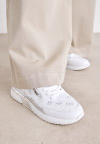 Le sneakers bianche presentano un design a motivi con sottili accenti grigi, pelle testurizzata e una suola a motivi, abbinate a pantaloni beige.