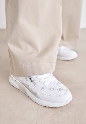 Le sneakers bianche presentano un design a motivi con sottili accenti grigi, pelle testurizzata e una suola a motivi, abbinate a pantaloni beige.