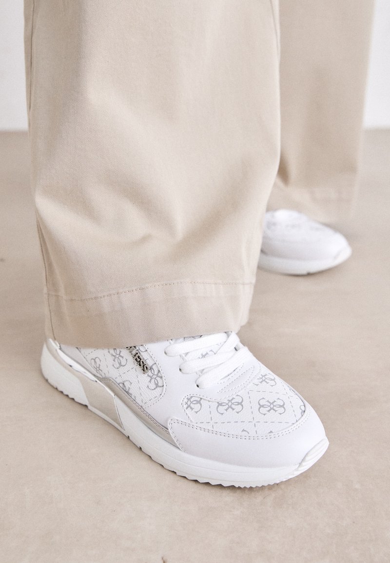 Le sneakers bianche presentano un design a motivi con sottili accenti grigi, pelle testurizzata e una suola a motivi, abbinate a pantaloni beige.