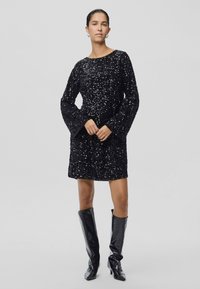 Kobieta w czarnej, długozasłonowej sukience z cekinami i czarnych, sięgających za kolano, teksturowanych botkach, pozująca na jednolitym jasnym tle.