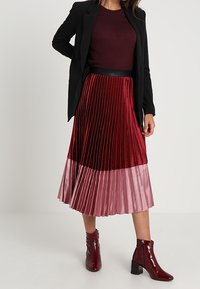 Rode geplooide rok met variërende tinten van donker bordeauxrood tot lichtroze, gedragen met een zwarte getailleerde blazer en bordeauxrode enkellaarsjes.