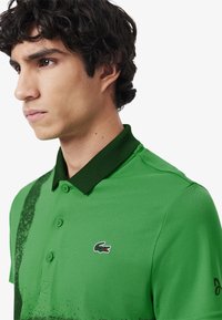 Grön polo skjorta med en texturerad yta, prydd med en mörkare grön krage och ett subtilt sprutmönster. Lacoste-logotypen på vänster bröst.