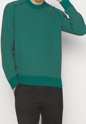 Mann trägt texturierten grünen Pullover mit langen Ärmeln und schwarze Hose, eine Hand in der Tasche und die andere in Kopfnähe erhoben.