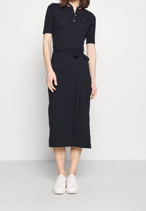 Kobieta ubrana w czarną midi sukienkę z krótkim rękawem i wiązanym paskiem w talii oraz białe trampki, stojąca na tle jednolitego jasnego tła.