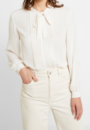 Femme portant un chemisier crème avec des plis et un nœud noué au niveau du col, associé à un pantalon en velours côtelé crème taille haute.