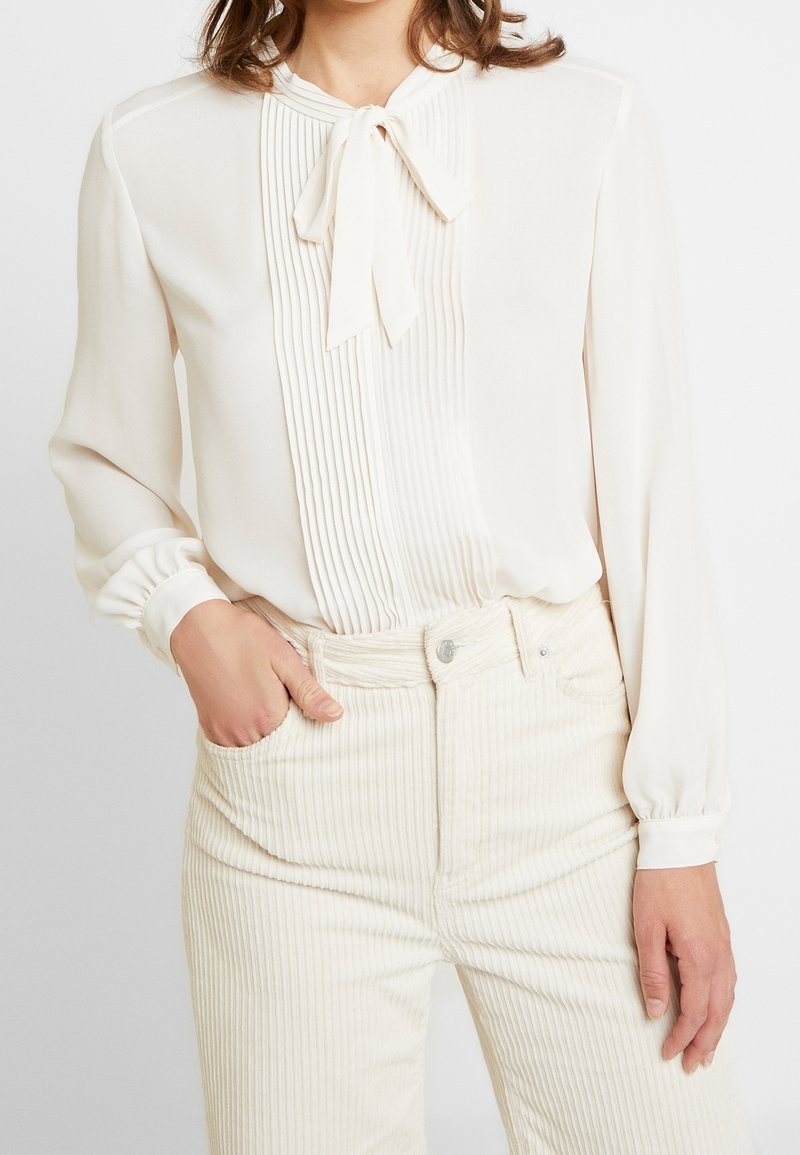 Femme portant un chemisier crème avec des plis et un nœud noué au niveau du col, associé à un pantalon en velours côtelé crème taille haute.
