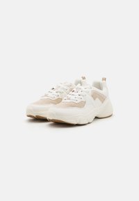 White sneakers with a textured beige mesh panel, rounded rubber sole, and flat laces. Features a contrasting beige accent on the sides.