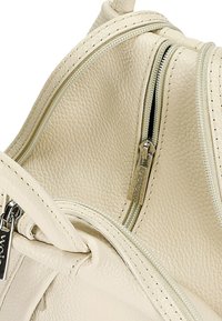 Krémová kožená kabelka s dvěma zipy, strukturovaným povrchem a viditelným stehem. Obsahuje kovový štítek s názvem značky.