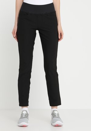 Frau trägt schwarze Slim-Fit-Hose mit elastischem Bund und hellgrauen Sportschuhen, steht vor einem schlichten weißen Hintergrund.