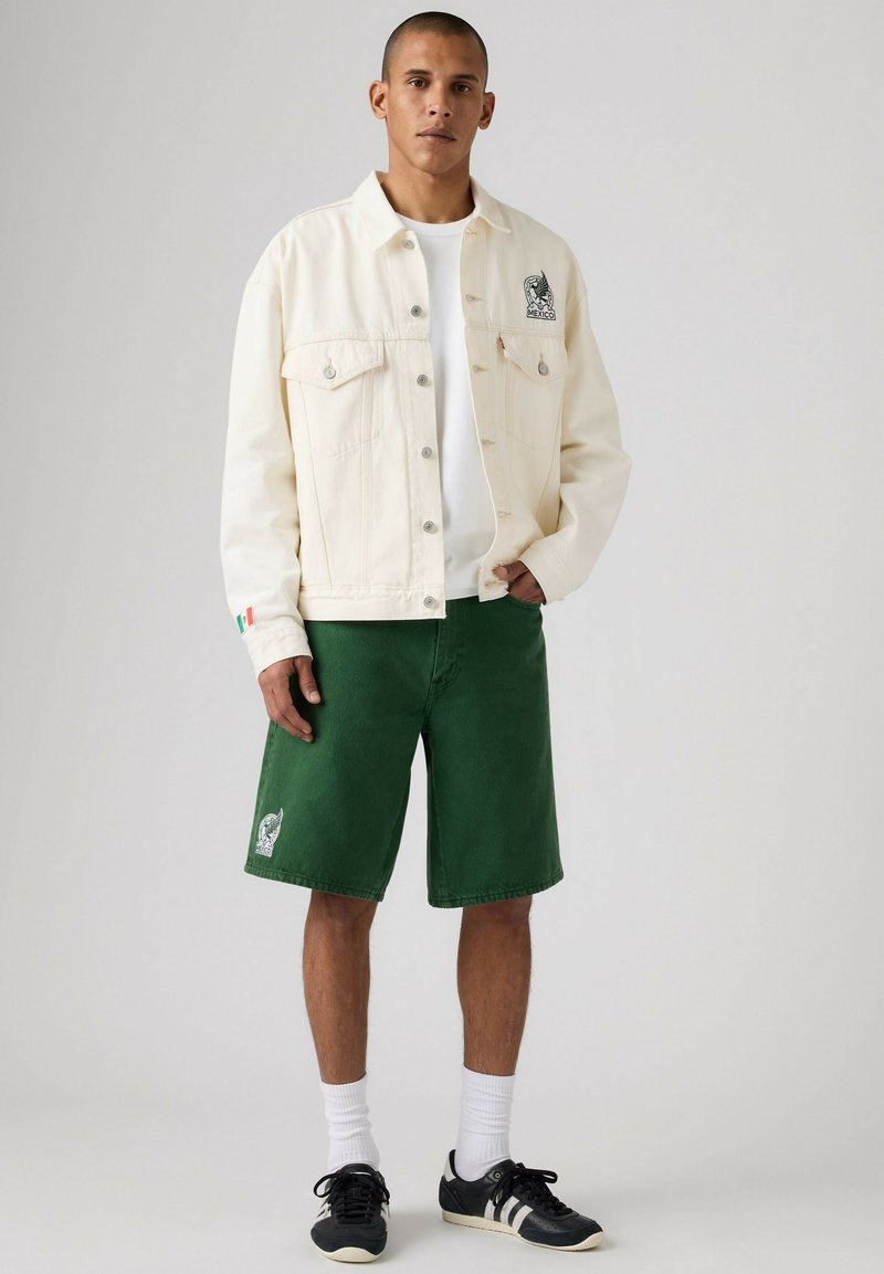 Ein Mann steht und trägt eine weiße Jacke mit Knöpfen und einem Mexiko-Emblem, grüne Shorts mit kleinem Logo, weiße Socken und schwarze Turnschuhe.