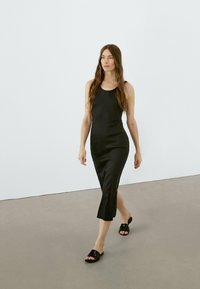 Abito midi senza maniche di colore nero con una silhouette aderente e uno spacco laterale, abbinato a sandali neri a scivolo. Tessuto liscio, design semplice.