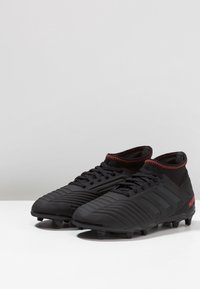 Black soccer cleats with a textured surface, high-top design, red accent stitching, and molded studs for traction.