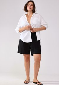 Camicia bianca in lino con bottoni e tasca, indossata parzialmente aperta su pantaloni corti neri con vita elastica. Infradito neri completano l'outfit.