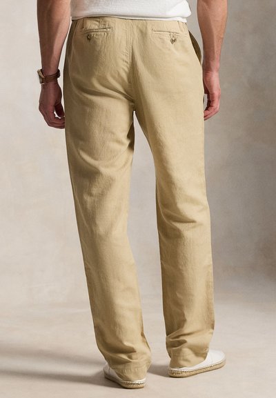 Άνδρας με μπεζ chinos και λευκά εσπαντρίγιες, που φαίνεται από πίσω, στέκεται μπροστά σε ουδέτερο φόντο.