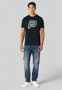 Juodas medvilnės marškinėliai su išskirtiniu baltu ir sidabriniu „P“ logotipu, derinami su mėlynomis džinsinėmis kelnėmis ir baltais sportiniais bateliais. Vienoda fono spalva.