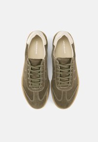 Παντοφλέ παπούτσια από πράσινο σουέντ με λευκό δερμάτινο γιακά, σχεδιασμό με κορδόνια, αντίθετες ραφές και σόλα από καουτσούκ χρώματος καραμέλας.