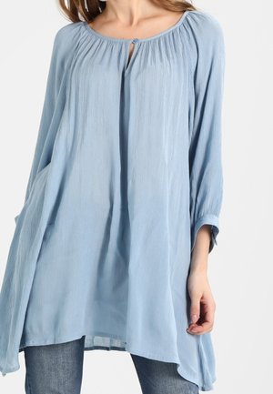 Femme portant une tunique ample bleu clair à manches longues avec un col froncé et un détail d'un seul bouton, associée à un jean bleu.