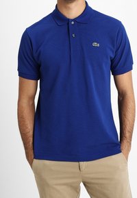 Polo shirt bleu royal en coton, avec un col classique, une patte de boutonnage à deux boutons, et un logo brodé vert sur la poitrine.