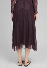 Jupe en dentelle de couleur violet profond avec un design superposé, une texture transparente et un ourlet fluide. Accompagnée de sandales brunes à lanières.