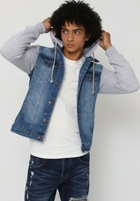 Gilet en denim avec une teinte bleue, manches de sweat à capuche grises, poches avant et boutons décoratifs. Porté par-dessus une chemise blanche à motif texturé.