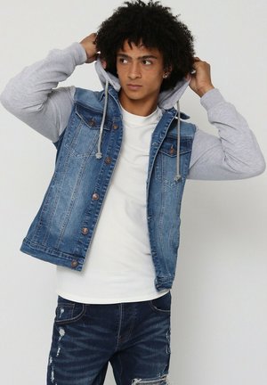 Gilet en denim avec une teinte bleue, manches de sweat à capuche grises, poches avant et boutons décoratifs. Porté par-dessus une chemise blanche à motif texturé.
