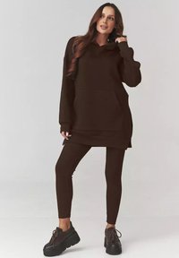 Brązowa, oversize'owa bluza z dużą kieszenią z przodu, połączona z brązowymi legginsami o prążkowanej fakturze oraz ciemnymi sneakersami na platformie. Gładka, miękka tekstura.