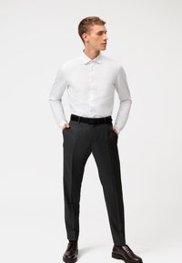 Chemise blanche à manches longues avec col boutonné, assortie à un pantalon tailleur gris foncé et une ceinture noire, portée avec des chaussures noires.