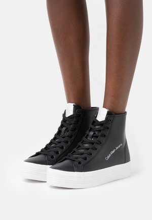 Zapatillas altas negras hechas de material sintético suave, con suelas de goma blanca y cordones, con la marca "Calvin Klein Jeans".