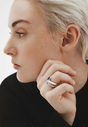 Anillo de plata con una piedra preciosa negra y banda texturizada, llevado en una mano apoyada contra el mentón. Fondo neutro, vista de perfil lateral del modelo.
