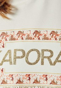 Witte shirt met gouden glanzende tekst "ZAPORA." Bevat een bloemenpatroon band in roze en bruin met gouden accenten.