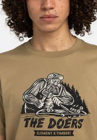 Krátký rukáv, pískově zbarvené tričko s černou grafikou vousatého muže, který opracovává dřevo, v pozadí stromy a text "DĚLAJÍCÍ".