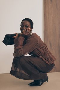 Brūna plisēta blūze ar gariem piedurknēm, ar viļņotu dibenu; caurspīdīga brūna svārku; melna rokassoma; melni norādīti papēži; gaiši koka fons.