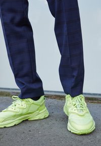 Kirkkaan neonkeltaiset lenkkarit teksturoidulla materiaalilla ja monimutkaisella kuviolla, yhdistettyinä tummansinisiin ruudullisiin housuihin, joissa on hienovaraiset viivat.