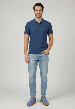 Lächelnder Mann steht mit einer Hand in der Tasche, trägt ein marineblaues Poloshirt, hellblaue ausgewaschene Jeans und blaue Sneakers mit braunen Sohlen.