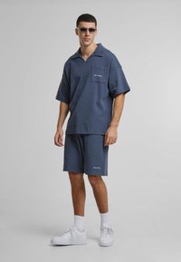 Un outfit casual pentru bărbați, care include un polo texturat, lejer, de un albastru închis, cu un buzunar, și şorturi asortate, completat cu adidași albi.