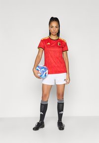 Κόκκινη ποδοσφαιρική φανέλα με κίτρινες λεπτομέρειες και έμβλημα του Βελγίου, λευκά σορτς, μαύρες κάλτσες με ρίγες, κρατώντας μια πολύχρωμη ποδοσφαιρική μπάλα.