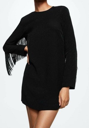 Frau trägt ein kurzes schwarzes Kleid mit langen Ärmeln und Fransenverzierung an einem Arm, steht mit einer Hand auf der Hüfte.