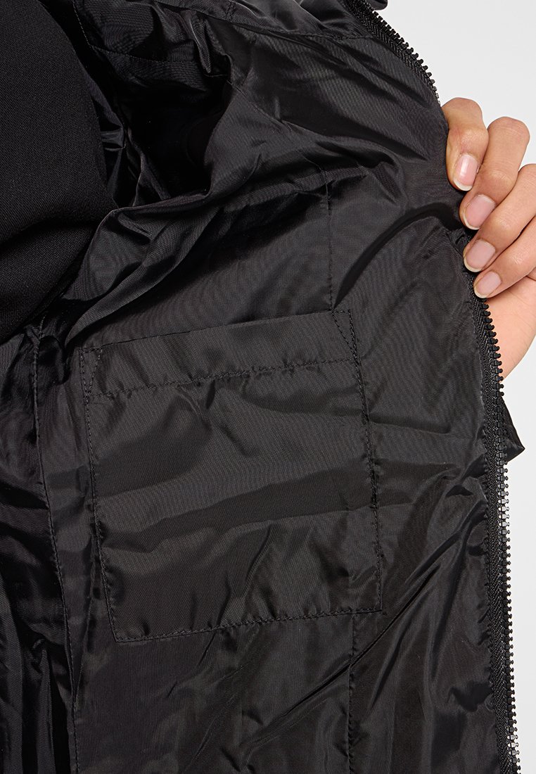 Mano sosteniendo una chaqueta negra abierta que muestra el suave tejido interior y un bolsillo rectangular cosido cerca del zipper.