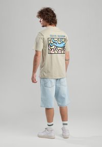 Camiseta de algodón beige con un gráfico colorido de Keith Haring en la parte posterior. Combinada con pantalones cortos de mezclilla azul claro y zapatillas blancas.