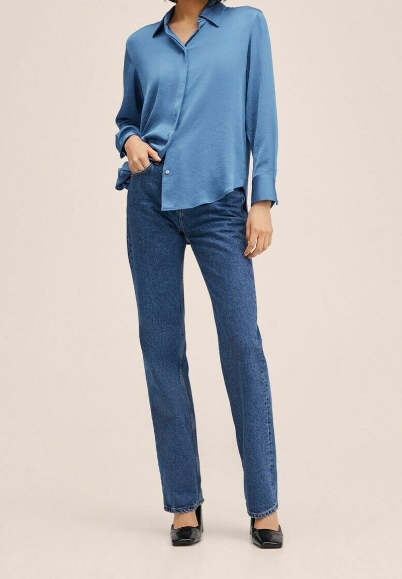 Femme portant une chemise bleue ample boutonnée, rentrée dans un jean bleu taille haute à jambes droites avec des chaussures noires à petit talon.