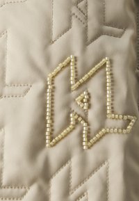 Béžová tkanina s prešívaným vzorom; zdobená dekoratívnym logom obrysom z malých, čírych korálkov; hladká textura, jemný lesk.