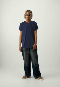 Jongeman met gevlochten haar, die donkere zonnebril draagt, een marineblauw t-shirt, losse donkere jeans en bruine sneakers aanheeft, staande op een lichte achtergrond.