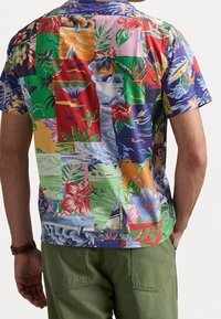 Uomo che indossa una camicia hawaiana a maniche corte multicolore con motivi floreali e barche a vela, abbinata a pantaloni verde oliva e un orologio da polso marrone.