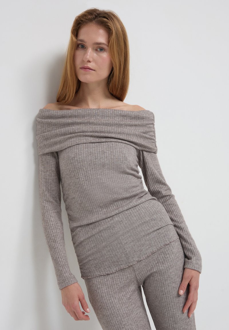 Junge Frau mit langen roten Haaren, die einen gerippten, schulterfreien beigen Pullover und passende Hosen trägt und vor einer weißen Wand steht.