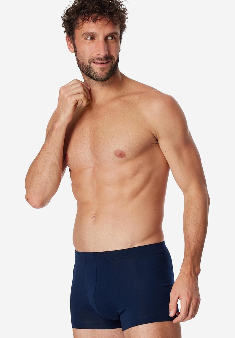 Homme aux cheveux bouclés et barbe portant un boxer bleu marine, debout contre un fond blanc, touchant son cou et regardant sur le côté.