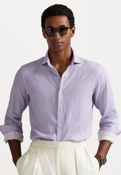 Camisa de botones de color púrpura claro con un patrón sutil, mangas arremangadas y puños blancos; combinada con pantalones plisados color crema y un reloj marrón.