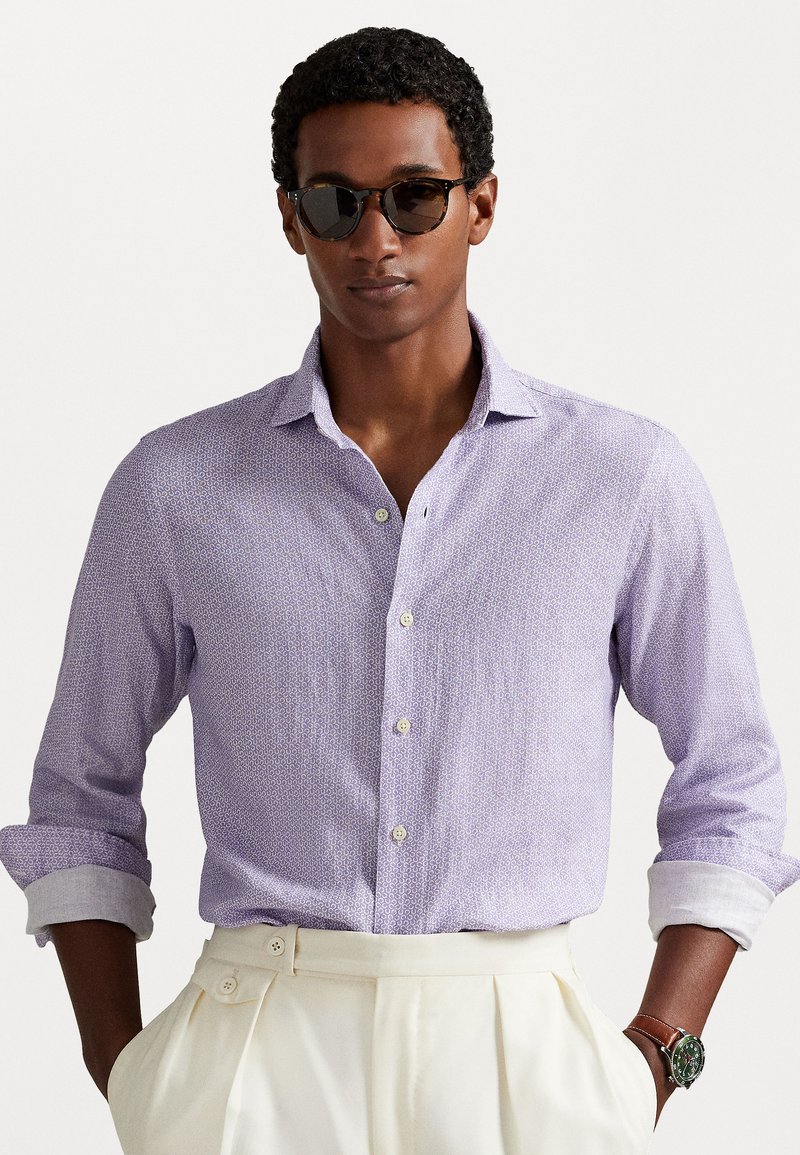 Chemise boutonnière lilas clair avec un motif subtil, manches retroussées et poignets blancs ; associée à un pantalon plissé crème et une montre marron.